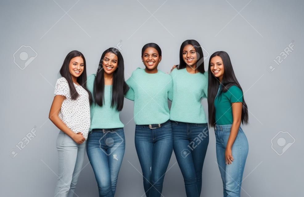 Group portrait of confident diverse women. Team of happy beautiful young black and white female friends in casual clothes standing together in studio, hugging each other, looking at camera and smiling