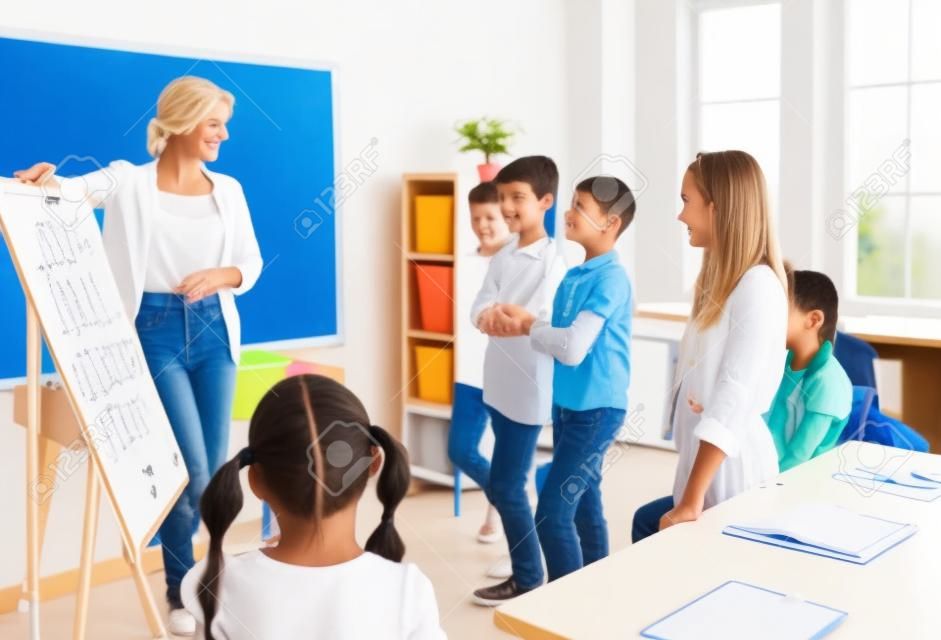 Happy teacher and students having interesting class in classroom. Educator uses white board, gives short presentation, explains grammar rules and asks children some questions. Back to school concept