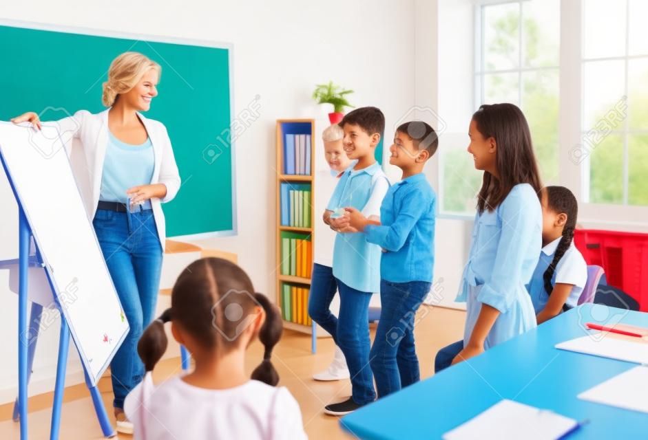 Happy teacher and students having interesting class in classroom. Educator uses white board, gives short presentation, explains grammar rules and asks children some questions. Back to school concept