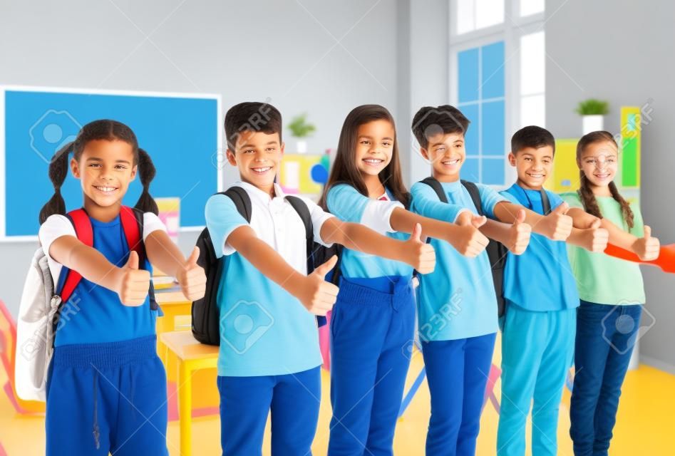 Groep leerlingen in het klaslokaal laten hun duimen zien om hun goedkeuring van grote keuze te tonen. Preteen jongens en meisjes met rugzakken op hun schouders staan op rij en glimlachen voor de camera.