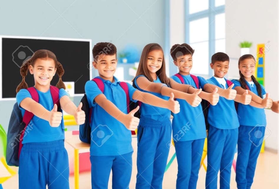Group of students in classroom show their thumbs up to show their approval of great choice. Preteen boys and girls with backpacks on their shoulders stand in row and smile at camera. Education concept