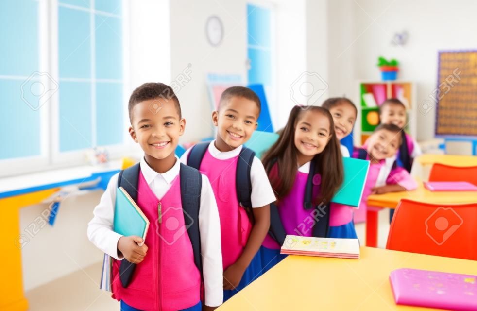 Onderwijs voor kinderen. Vrolijke schattige basisschool studenten poseren in het klaslokaal op 1 september. Kleine jongens en meisjes met boeken en rugzakken staan een achter elkaar en glimlachen voor de camera.