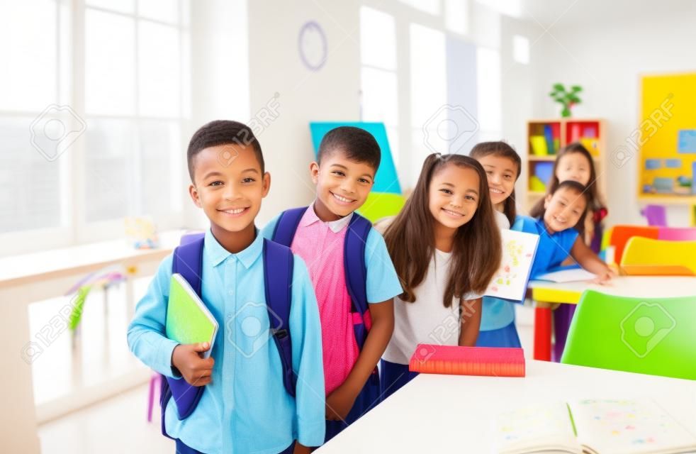 Educação para crianças. Estudantes de escola primária bonitos alegres posam em sala de aula no primeiro de setembro. Meninos e meninas com livros e mochilas ficam um atrás do outro e sorriem para a câmera.