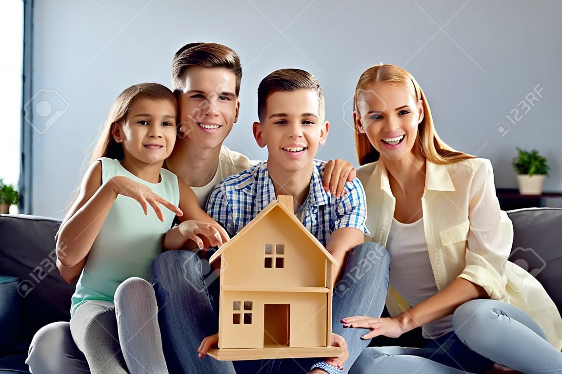 Portrait d'une jeune famille caucasienne heureuse avec deux enfants emménageant ensemble dans leur nouvel appartement lumineux. la famille est assise avec une petite maison en bois entre les mains. concept immobilier et logement familial.