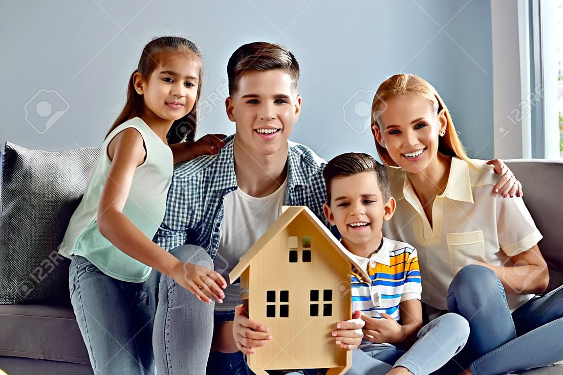Portret szczęśliwej młodej kaukaskiej rodziny z dwójką dzieci wprowadzających się razem do nowego jasnego mieszkania. rodzina siedzi z małym drewnianym domem w rękach. koncepcja nieruchomości i mieszkań rodzinnych.