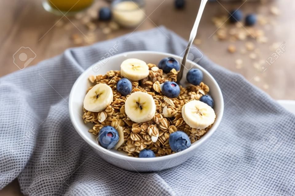 Pouring, adding milk to homemade granola in a plate with nuts, honey, blueberries and banana, served on napkin