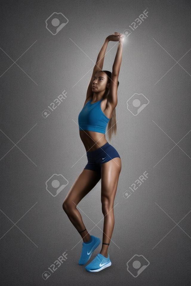 Full length shot of a young athletic slim mixed race fitness woman in sportswear stretching her arms, warming up while standing isolated over grey background