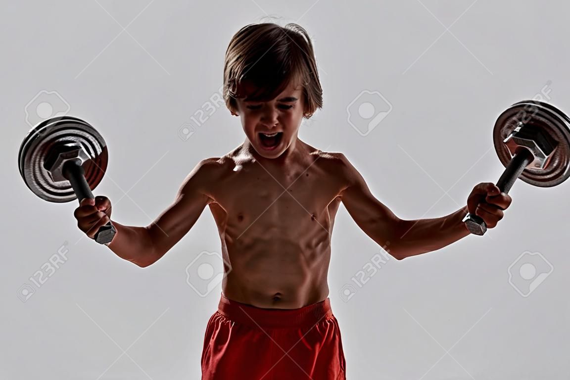 Kleiner sportlicher Junge mit muskulösem Körper, der beim Training emotional aussieht, Gewichte hebt, isoliert vor grauem Hintergrund steht