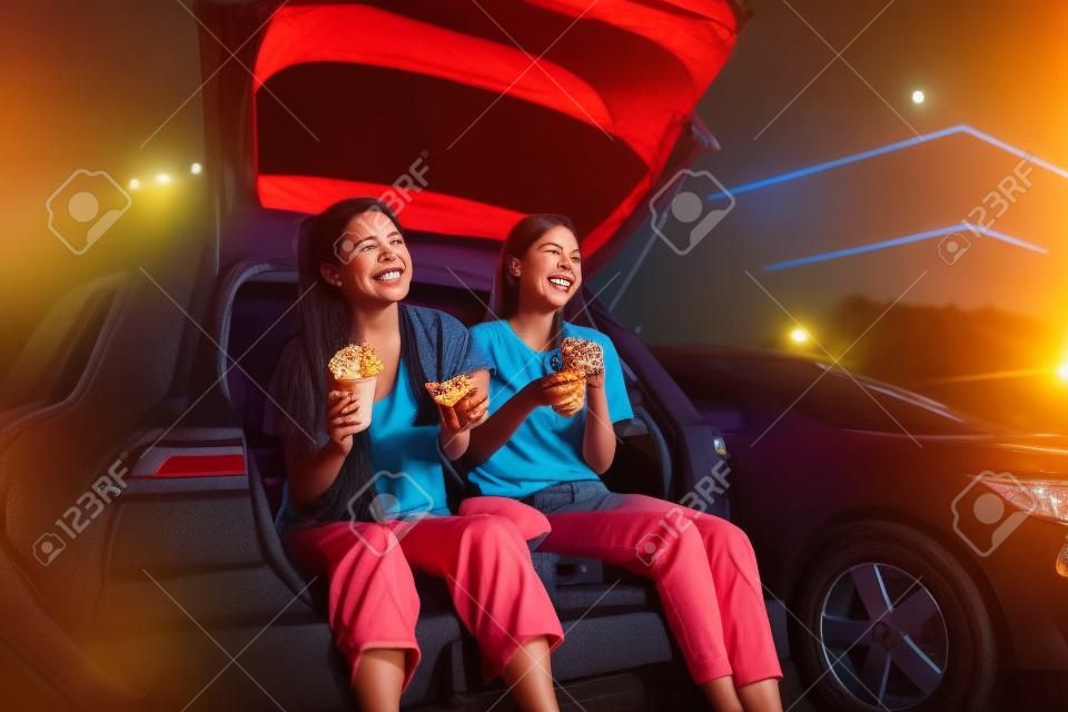 Two happy female best friends smiling while spending time together, having snacks and watching a movie in an outdoor cinema in the evening