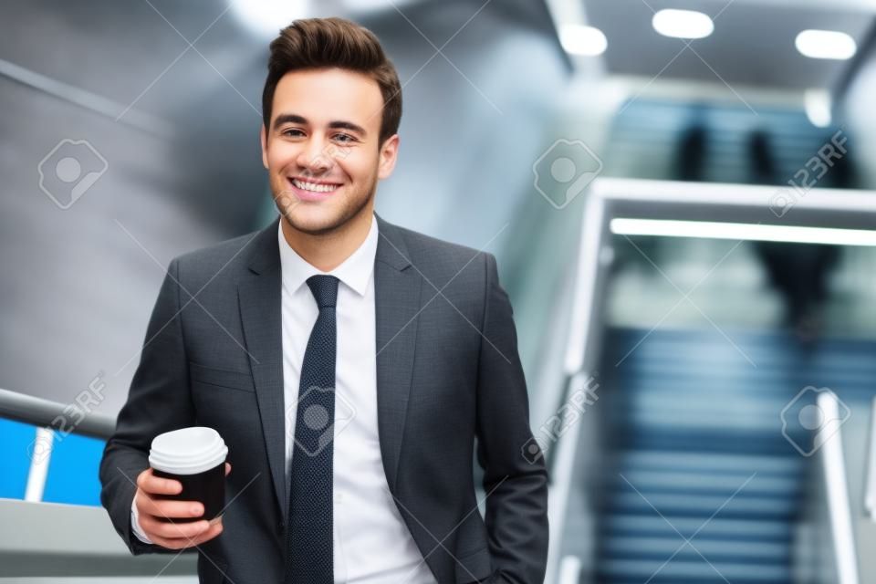 Smiling young executive in a suit drinking a coffee and riding up an escalator in a subway station during his morning commute