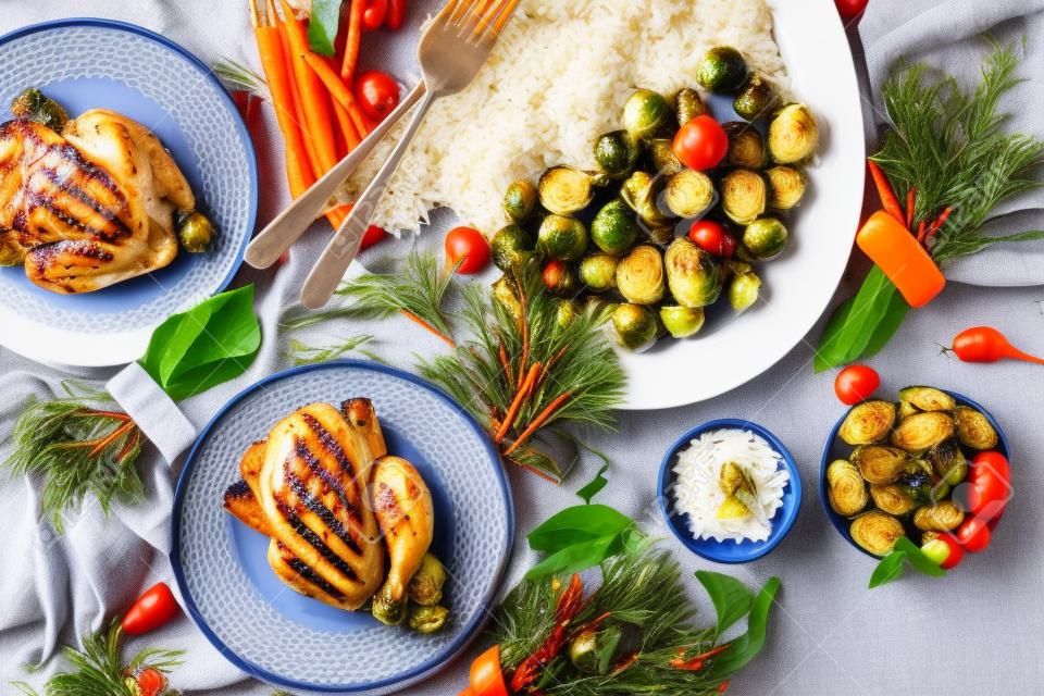 Weihnachtstisch mit gegrilltem Mini-Huhn, Reis und Gemüse, gebackener Rosenkohl, Babykarotten in Keramikschalen, Weihnachtsdekorationen auf blauem Leinentischtuch auf grauem Hintergrund. Flach liegen.