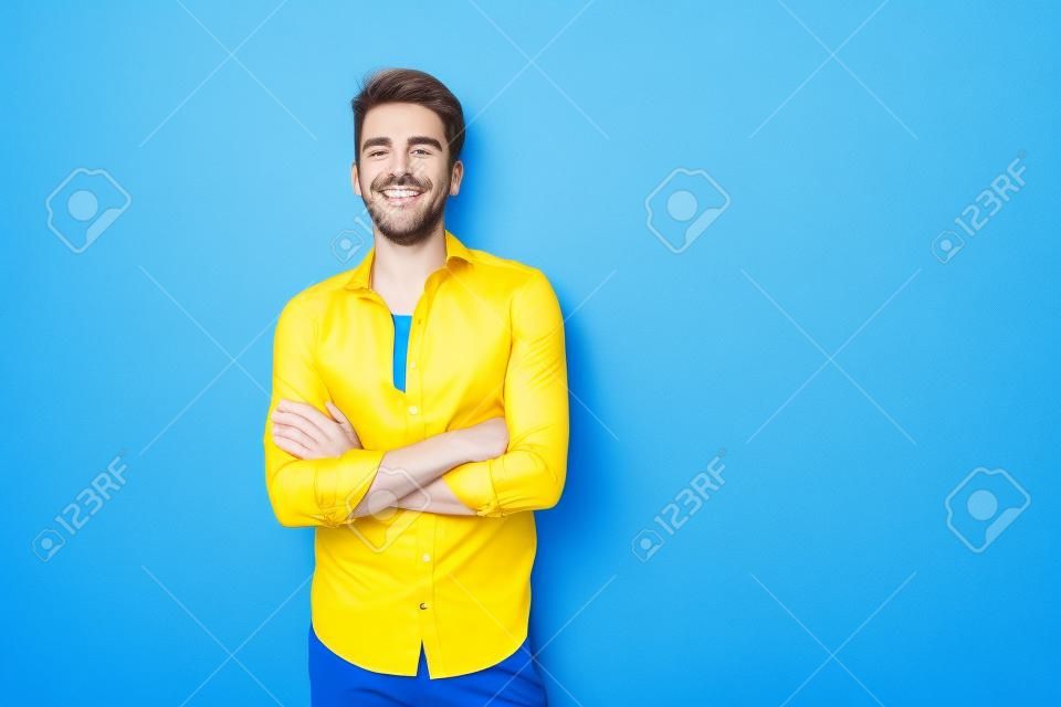 Vriendelijk uitziende vrolijke Europese man draagt blauw shirt glimlachend staand over gele achtergrond met gekruiste handen