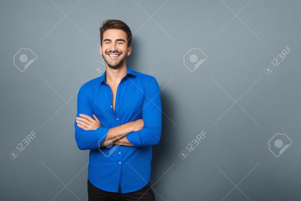 Un tipo europeo alegre y de aspecto amistoso con camisa azul sonriendo de pie sobre un fondo amarillo con las manos cruzadas