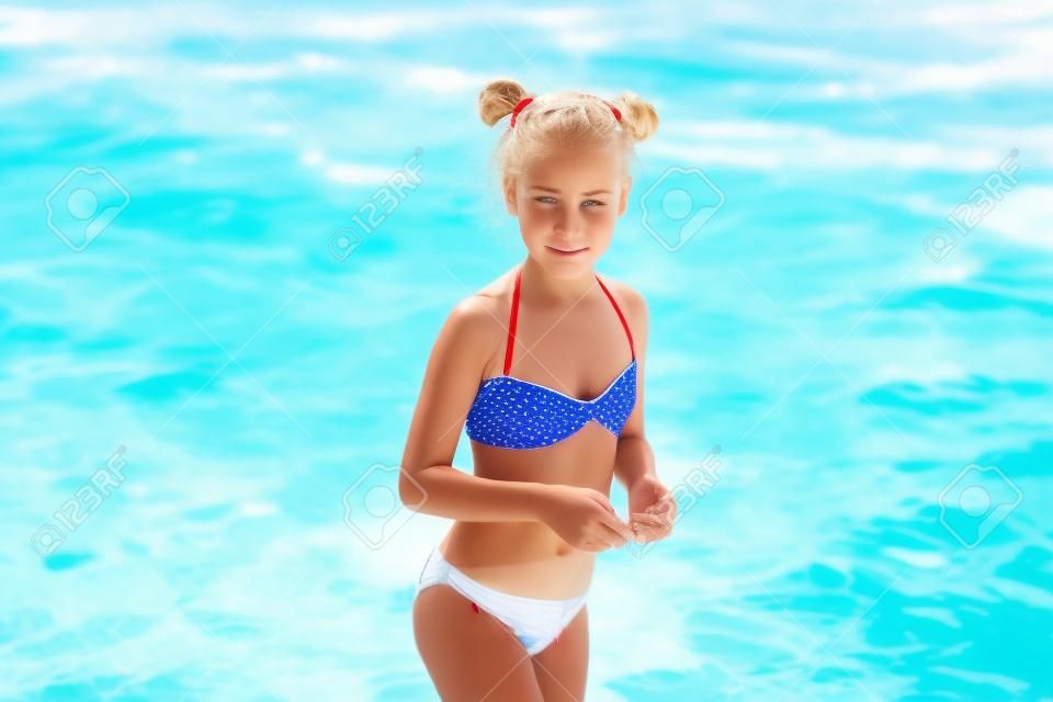 Schattig ontevreden blond meisje in het witte, rode en blauwe zwempak op strandvakantie. Blauwe zee met witte golven op de achtergrond.