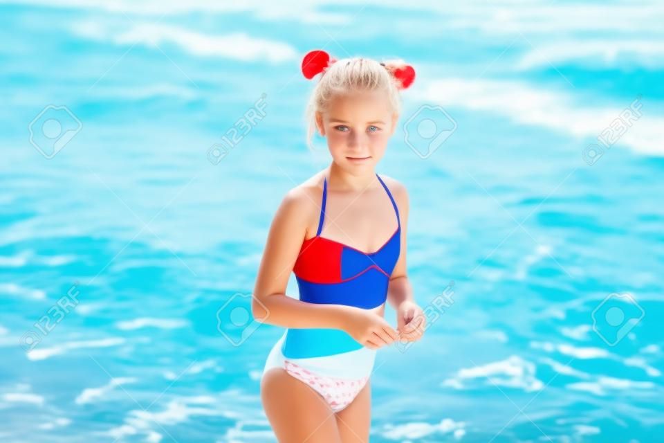 Adorable chica rubia insatisfecha con traje de baño blanco, rojo y azul en vacaciones en la playa. Mar azul con olas blancas en el fondo.