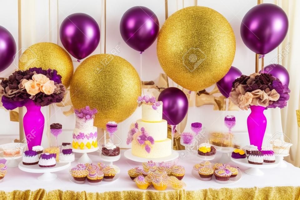다양한 사탕, 컵케이크, 수플레, 케이크가 있는 황금 결혼식 파티의 캔디 바. 갈색과 보라색, 자연 및 에코 테마로 장식된 실내입니다.
