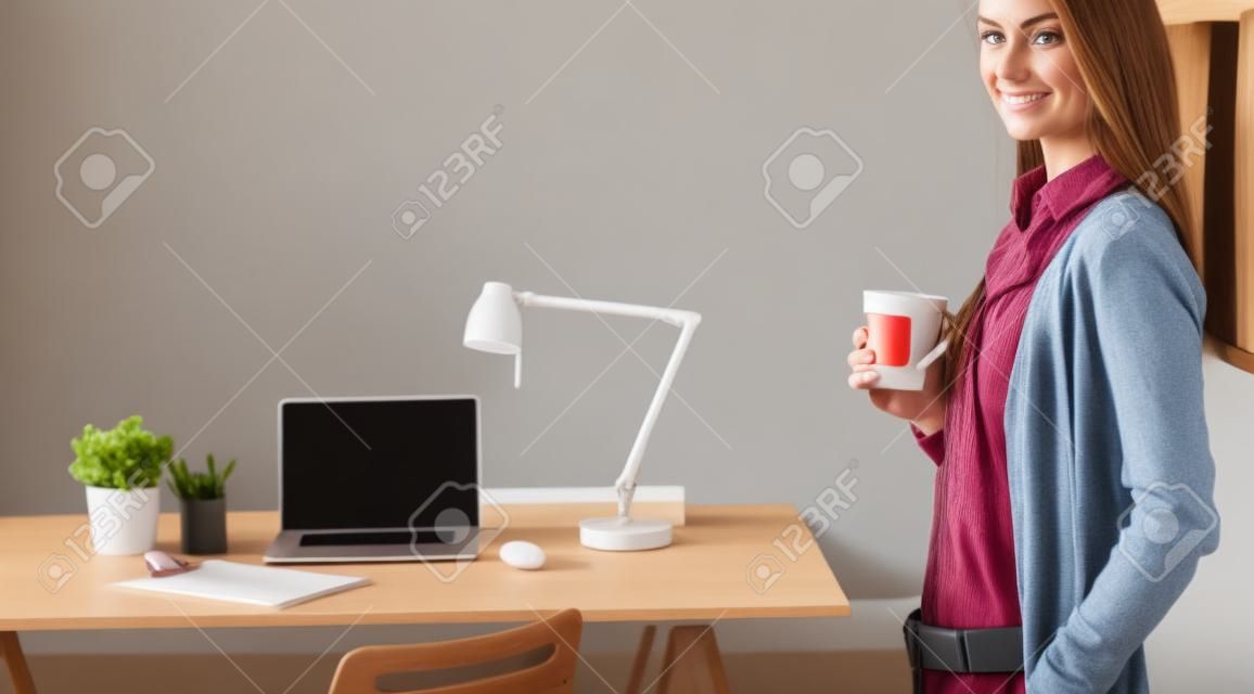 Portret zrelaksowanej młodej kobiety siedzącej przy biurku i trzymającej filiżankę kawy