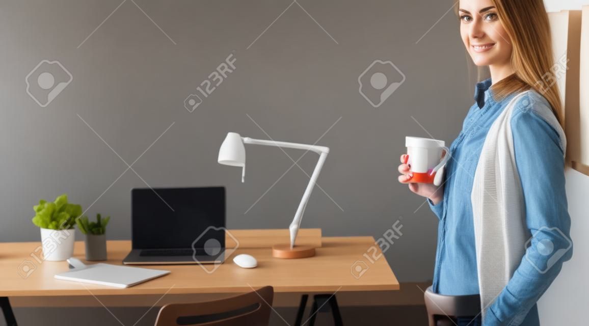 Portret zrelaksowanej młodej kobiety siedzącej przy biurku i trzymającej filiżankę kawy