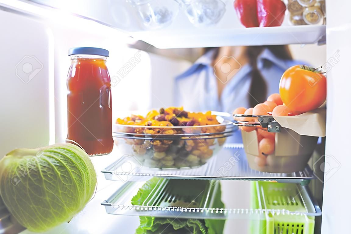 Retrato da fêmea que está perto da geladeira aberta cheia de alimento saudável, legumes e frutas.