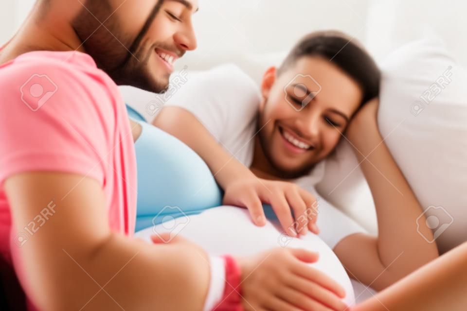 Young pregnant couple relaxing on sofa at home touching belly