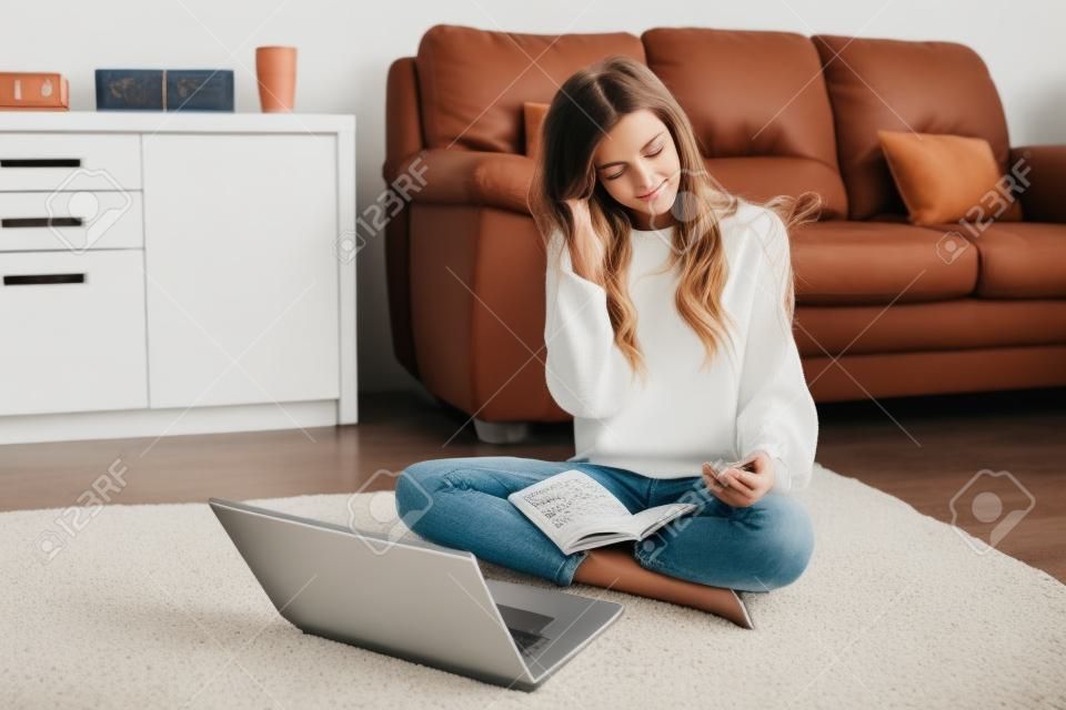 Charming young girl in a white pullover and jeans sitting on the floor with notebook.