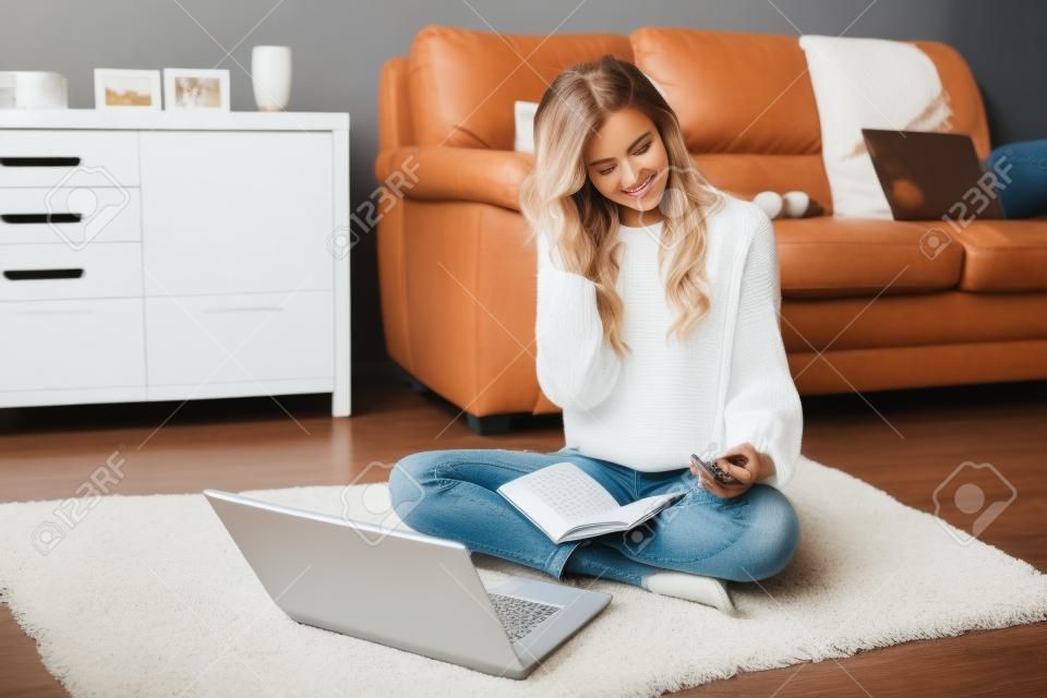 Charming young girl in a white pullover and jeans sitting on the floor with notebook.