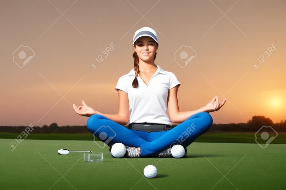 Mädchengolfer, der in Yogahaltung auf Golfplatz sitzt.