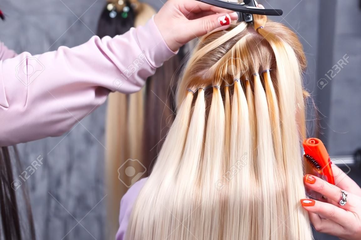 Procedimiento de extensiones de cabello. El peluquero hace extensiones del pelo a la muchacha joven, rubia en un salón de belleza. Enfoque selectivo.