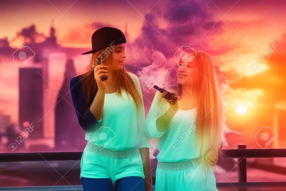 Two women vaping outdoor. The evening sunset over the city. Toned image.