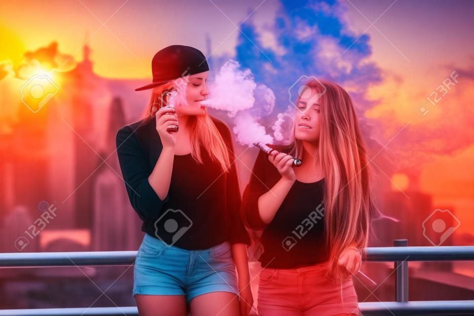 Two women vaping outdoor. The evening sunset over the city. Toned image.