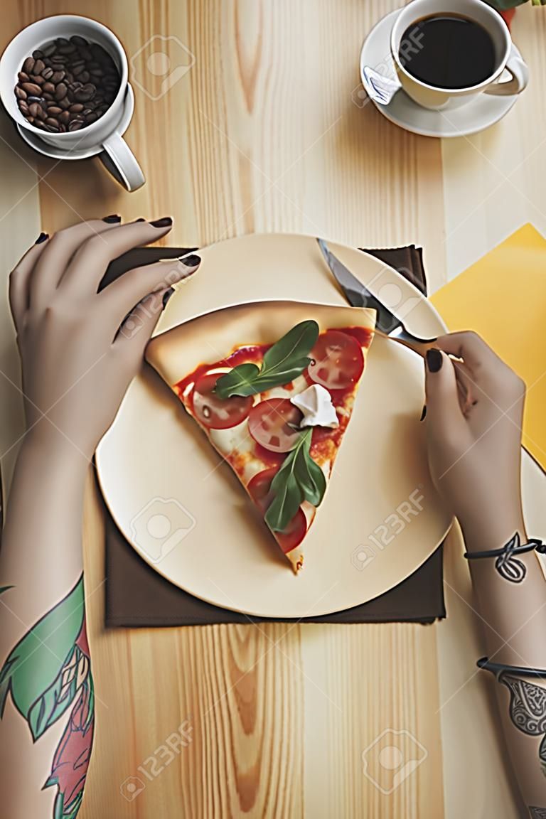 partial view of woman sitting at table with pizza piece on plate and cup of coffee