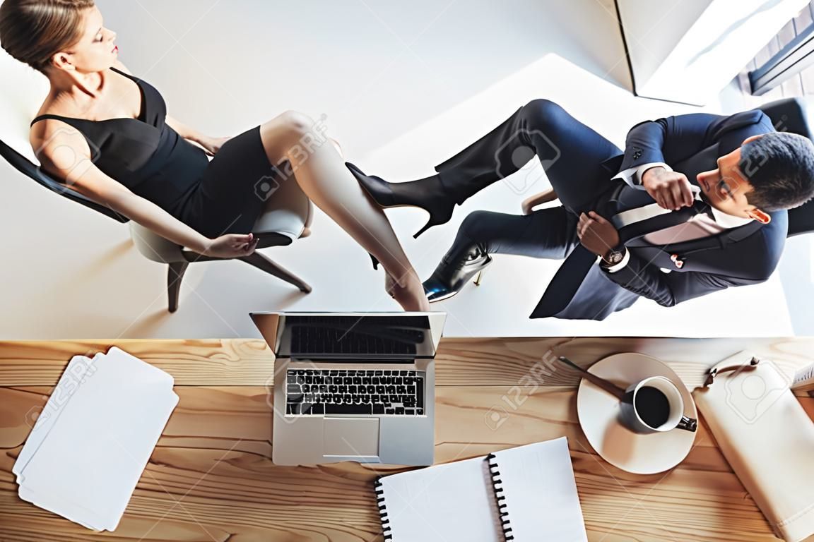 Vrouw in zwarte jurk en hoge hakken zet haar voet op een stoel tussen de benen van jongeman in zakenpak
