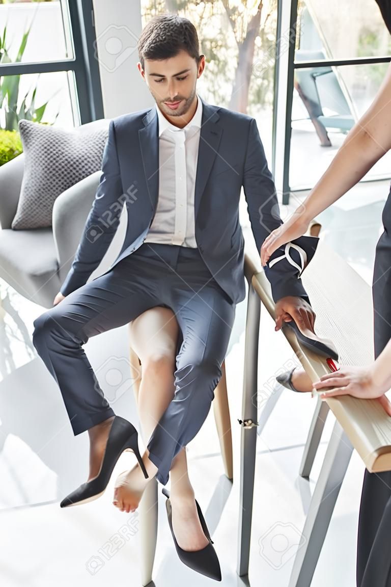 Женщина на высоких каблуках ставит ногу на стул между ног молодого человека в деловом костюме