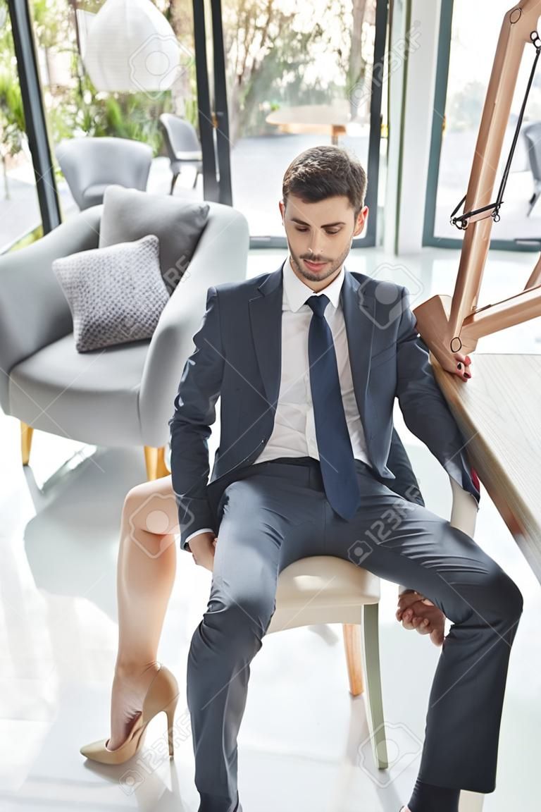 Женщина на высоких каблуках ставит ногу на стул между ног молодого человека в деловом костюме