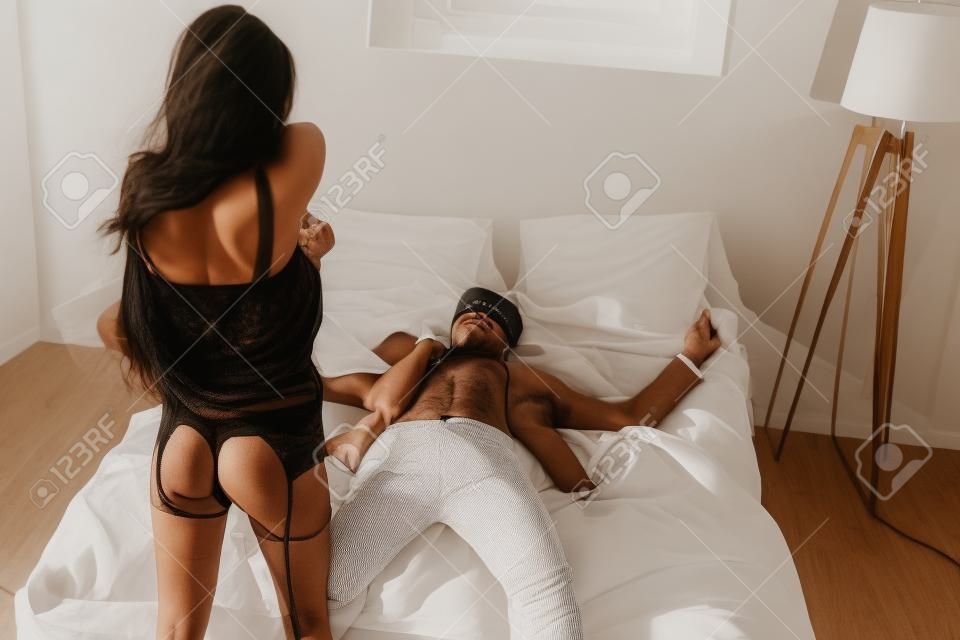 Frau zieht sich aus, während ihr Freund mit verbundenen Augen im Bett liegt