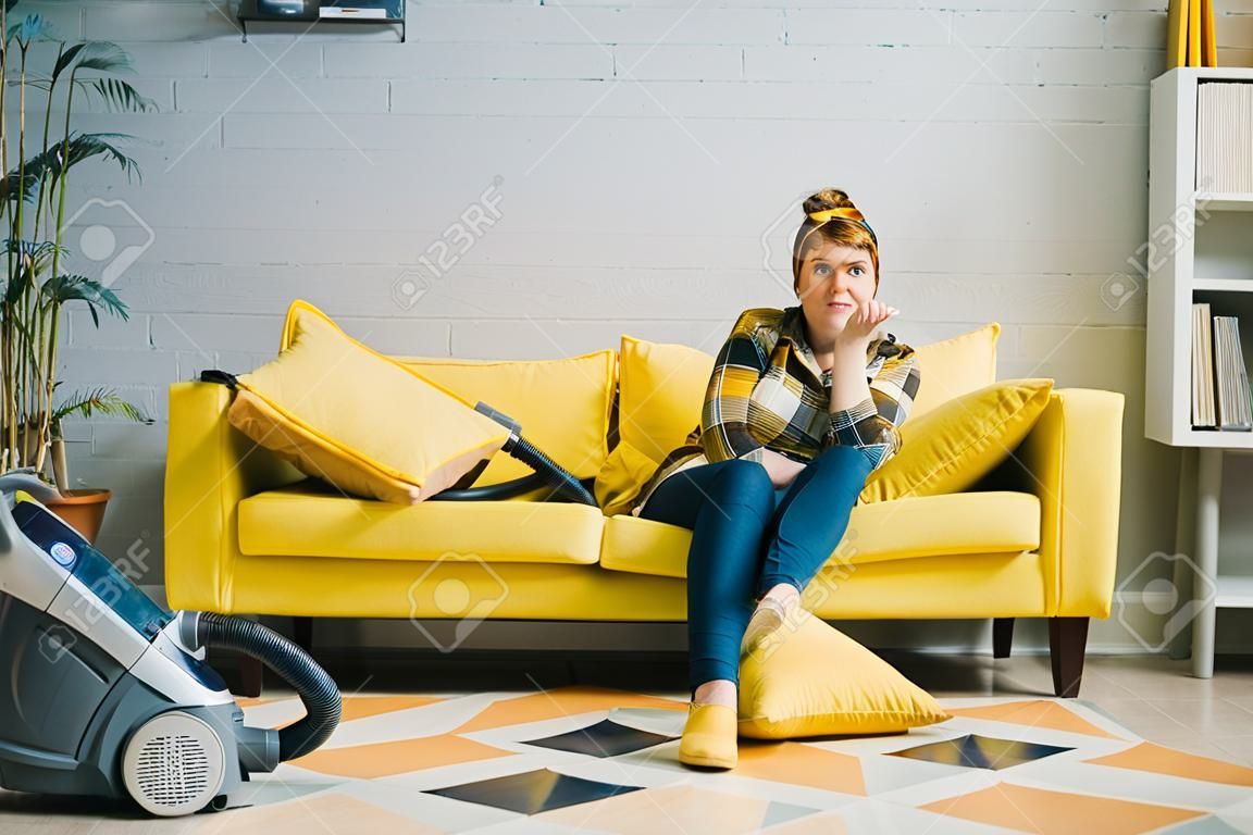 grimacing woman sitting on sofa and looking at camera, vacuum cleaner on floor at home