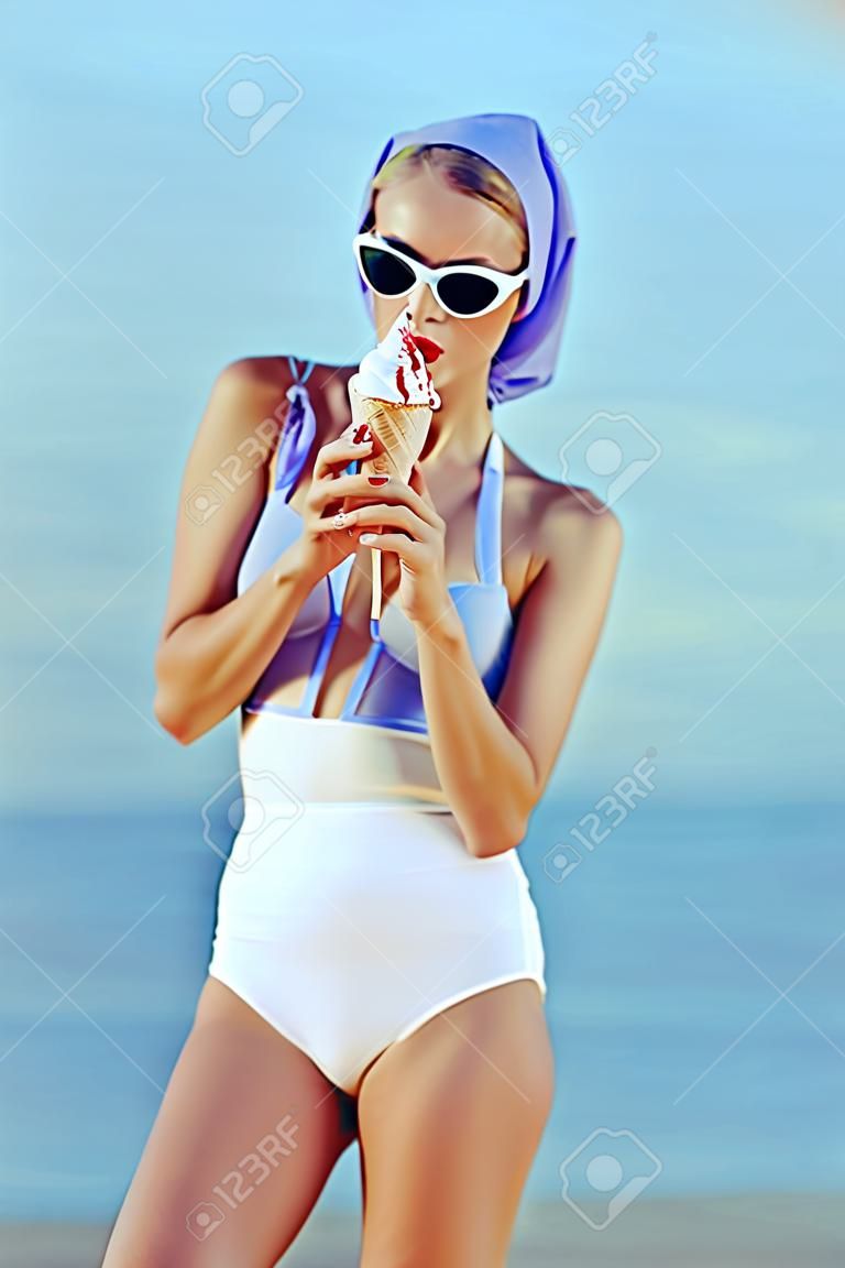 아이스크림을 들고 바다 근처에서 포즈를 취하는 빈티지 수영복과 선글라스를 쓴 우아한 소녀