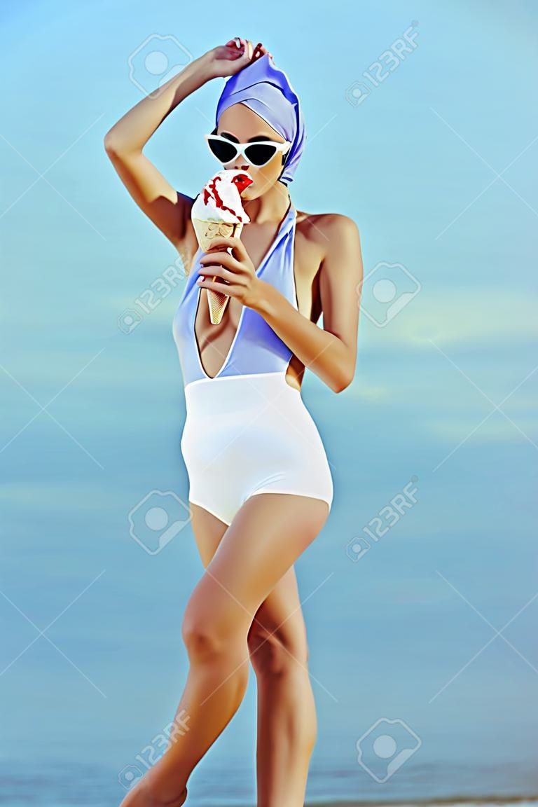 老式的泳装和太阳镜，拿着冰淇淋，并在海边摆姿势的优雅女孩