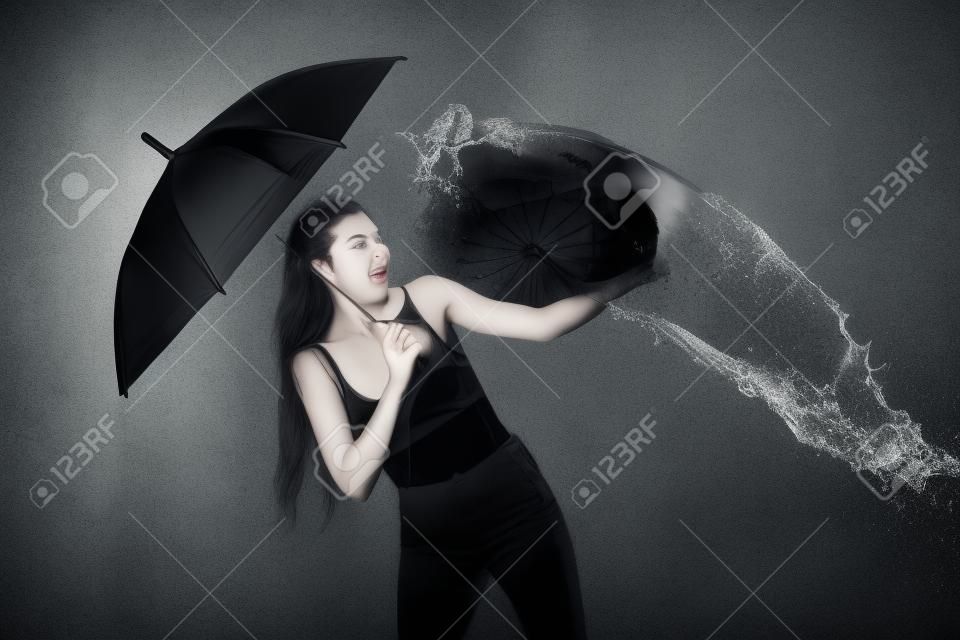молодая эмоциональная женщина с зонтиком, наполненная водой, изолированная на черном