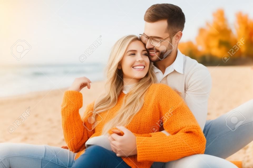 Smiling girlfriend and boyfriend in autumn outfit sitting and hugging on beach