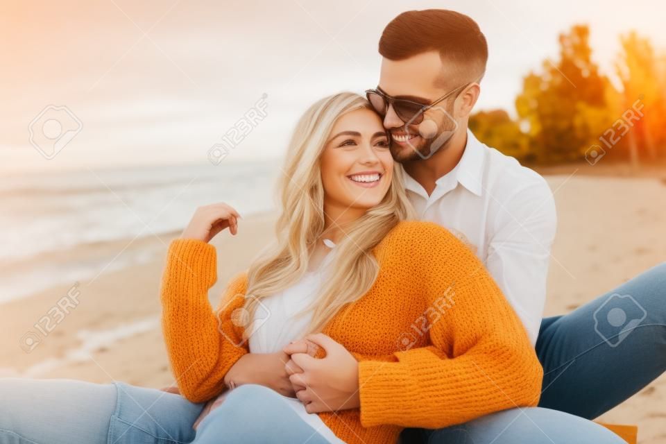 Lächelnde Freundin und Freund im Herbstoutfit sitzen und umarmen sich am Strand