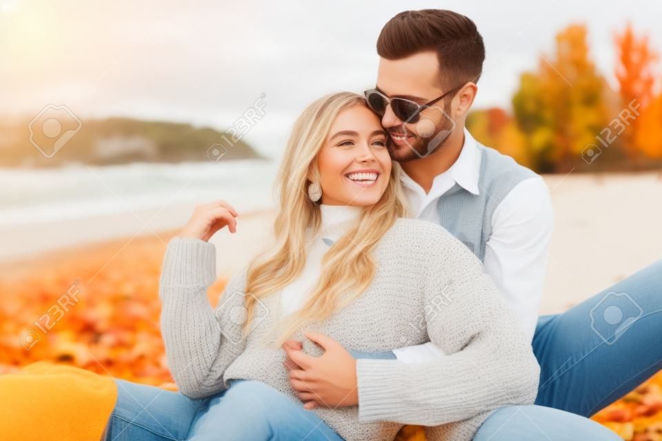 해변에 앉아서 포옹하는 가을 옷을 입은 웃는 여자 친구와 남자 친구