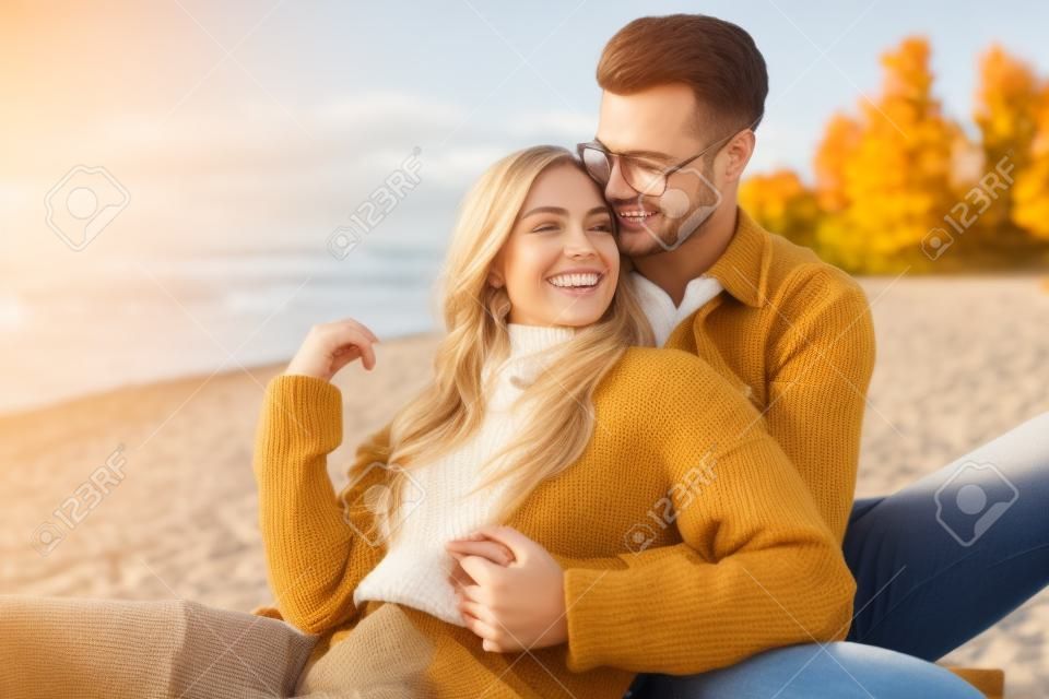 Sonriente novia y novio en traje de otoño sentado y abrazándose en la playa