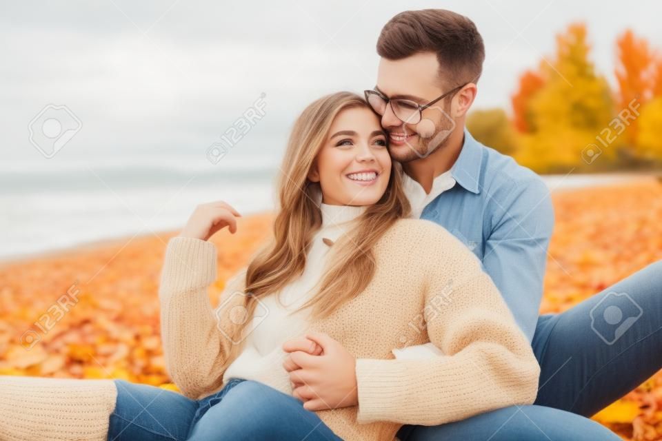 Uśmiechnięta dziewczyna i chłopak w jesiennym stroju siedzą i przytulają się na plaży