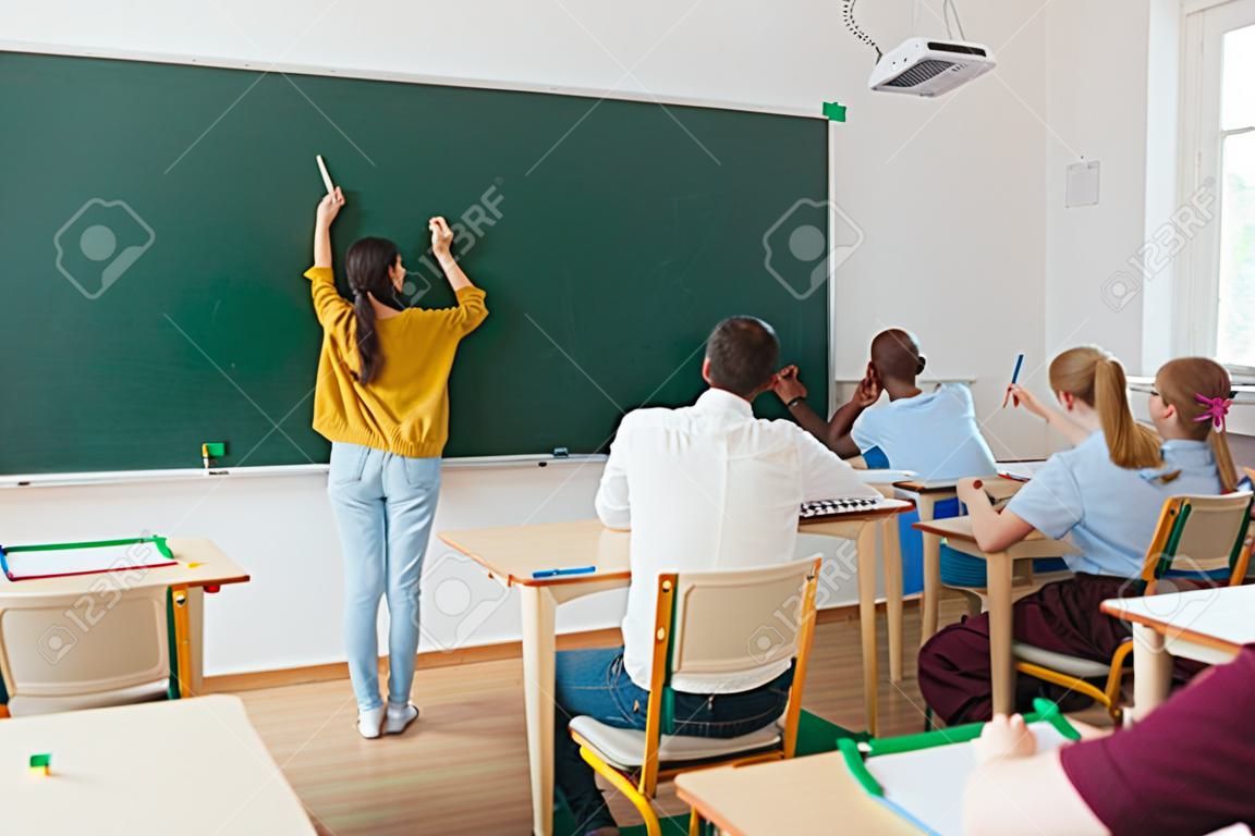Vista trasera de la colegiala escribiendo en la pizarra y compañeros de clase con el maestro sentado en el aula