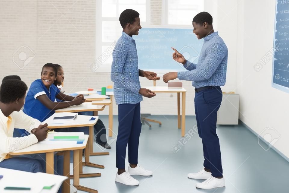 Side view of African American teacher pointing at wristwatch to latecomer schoolboy in classroom