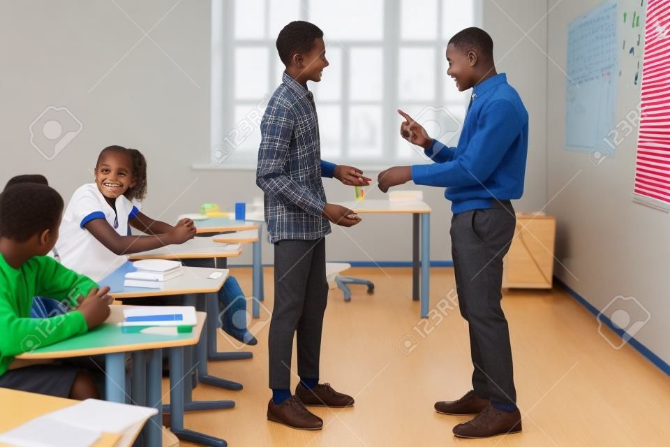 Side view of African American teacher pointing at wristwatch to latecomer schoolboy in classroom