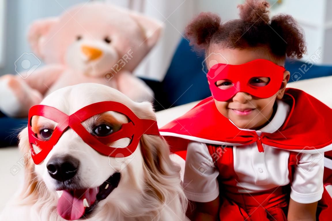 Smiling adorable little african american kid with happy dog in red superhero mask