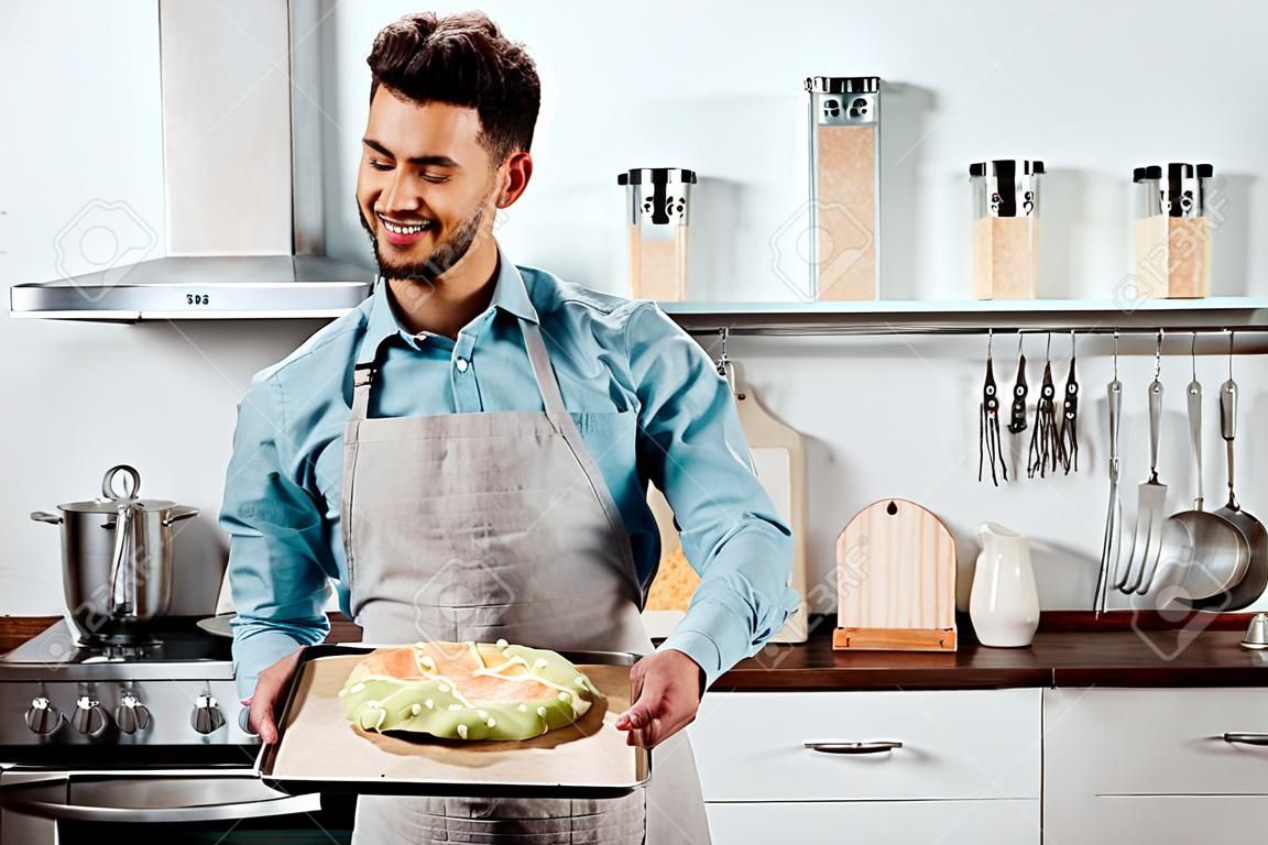 smiling young man in apron and potholders holding baking tray with fresh homemade pizza