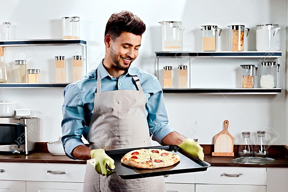 smiling young man in apron and potholders holding baking tray with fresh homemade pizza
